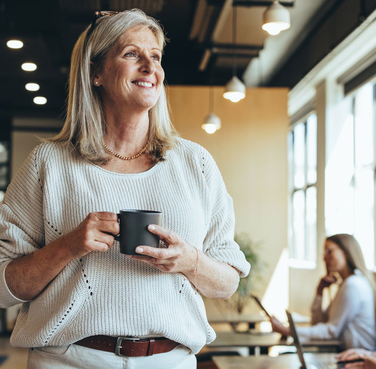 En leende äldre kvinna med grått hår, klädd i en mysig tröja, håller en kaffekopp medan hon står i en modern coworking-miljö. I bakgrunden arbetar yrkesverksamma på bärbara datorer, vilket skapar en dynamisk och samarbetsinriktad atmosfär.