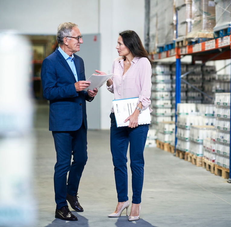 Två affärsproffs diskuterar logistik i ett lager och håller dokument och en surfplatta.