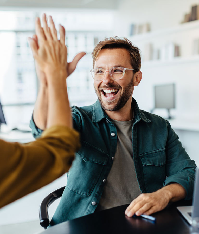 Ett glatt team på ett modernt kontor firar en framgång när en man och en kvinna gör en high-five. En annan kollega ler medan hon arbetar på sin laptop, vilket skapar en positiv och samarbetsinriktad arbetsmiljö.