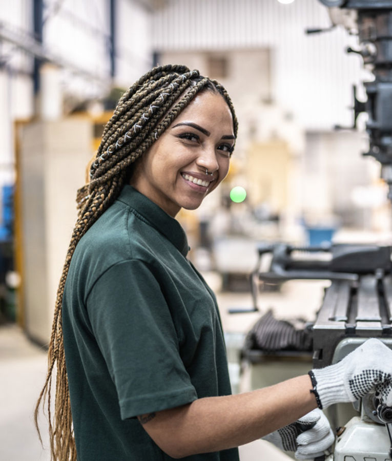 En leende kvinnlig maskinoperatör i mörkgrön uniform och handskar arbetar vid en fräsmaskin i en tillverkningsverkstad.