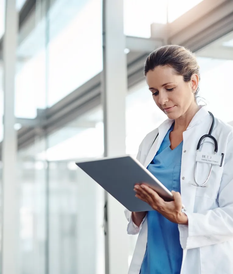 En kvinnlig läkare i vit rock med stetoskop granskar noggrant patientdata på en surfplatta i en ljus, modern sjukhuskorridor.