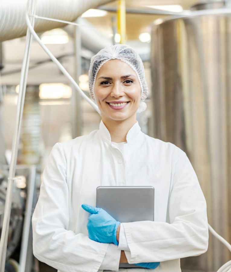 En leende kvinnlig kvalitetskontrollspecialist i vit labbrock, hårnät och handskar håller en surfplatta i en modern livsmedelsproduktionsanläggning.