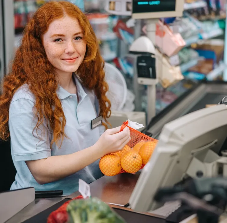 Leende ung kassörska i matbutik skannar apelsiner – symboliserar effektivitet, kassaarbete och kundbemötande.