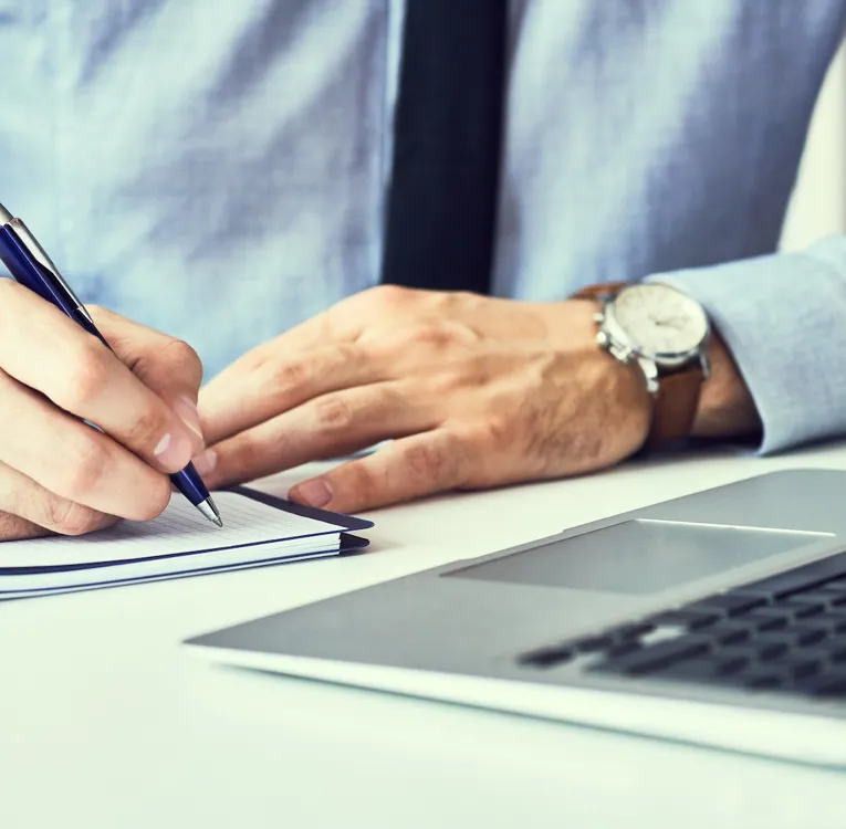 En närbild av en affärsman i en blå skjorta och slips som skriver anteckningar i en anteckningsbok medan han arbetar på en bärbar dator vid ett modernt skrivbord.