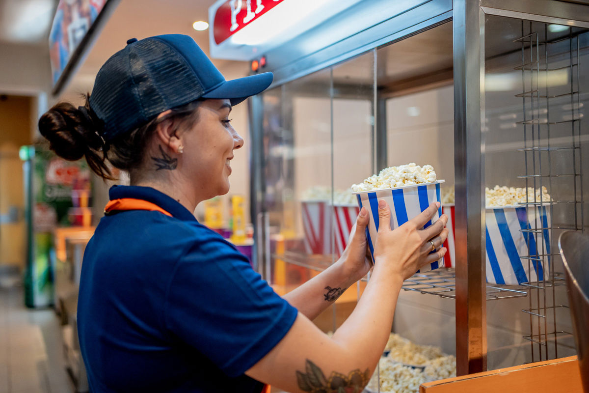 Leende biografanställd i uniform placerar en randig popcornbägare i ett värmeskåp – symboliserar skiftarbete, detaljservice och effektiv personalhantering.
