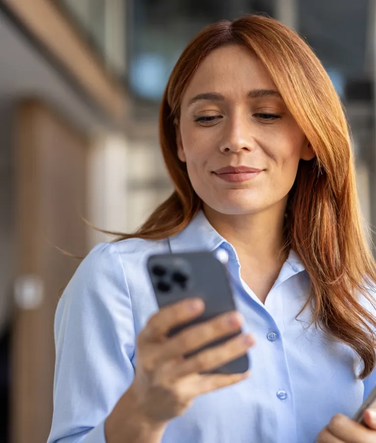Självsäker affärskvinna använder en smartphone medan hon håller en surfplatta och hanterar uppgifter på distans. Symboliserar mobil personalhantering och digital effektivitet.