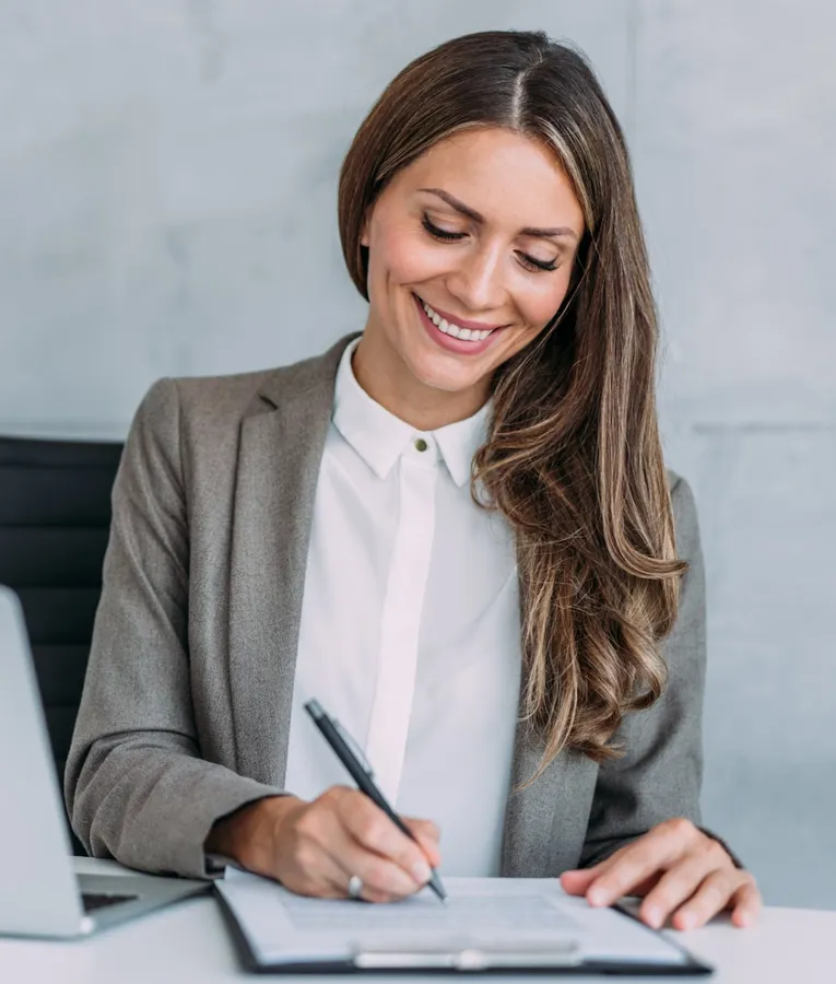 En leende affärskvinna i grå kavaj skriver under dokument vid sitt skrivbord med en bärbar dator bredvid.