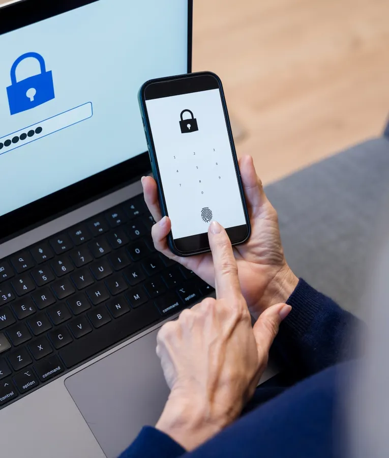 En person använder biometrisk autentisering på en smartphone medan de loggar in på en bärbar dator med en lösenordsskärm som visar en säkerhetsikon med lås.
