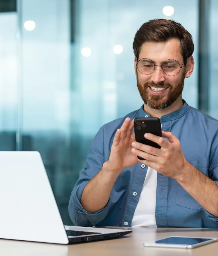 Leende affärsman använder en smartphone vid sitt skrivbord med en bärbar dator och surfplatta, som visar mobil arbetsstyrning på ett modernt kontor.