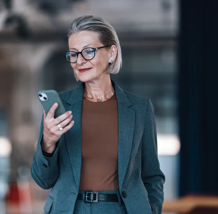 Självsäker senior affärskvinna använder en smartphone på ett modernt kontor för att hantera uppgifter och personalhantering på distans. Symboliserar ledarskap och mobil produktivitet.