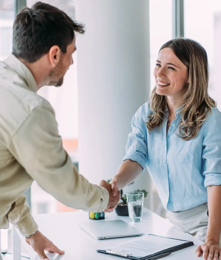 Leende affärskollegor skakar hand över ett skrivbord i ett ljust kontor – symboliserar lyckat samarbete, onboarding och en positiv arbetskultur.