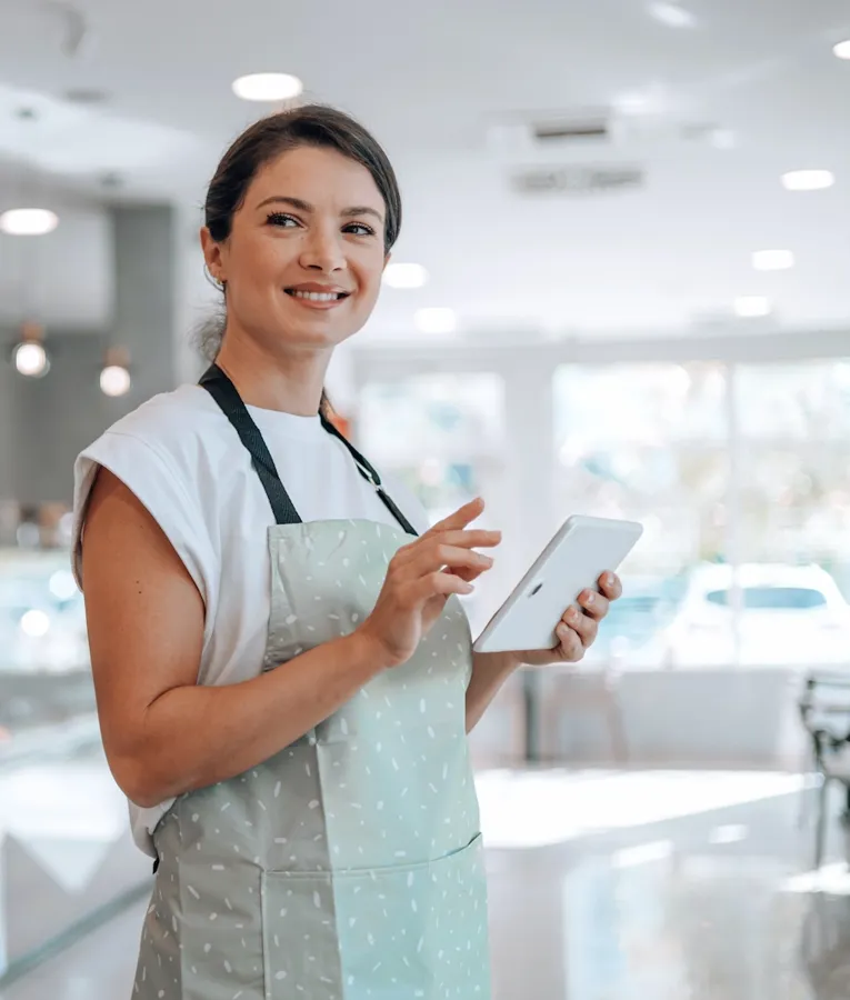 Leende bagerianställd använder en surfplatta för lager- eller orderhantering i ett modernt café, vilket visar digitala arbetsstyrningslösningar.