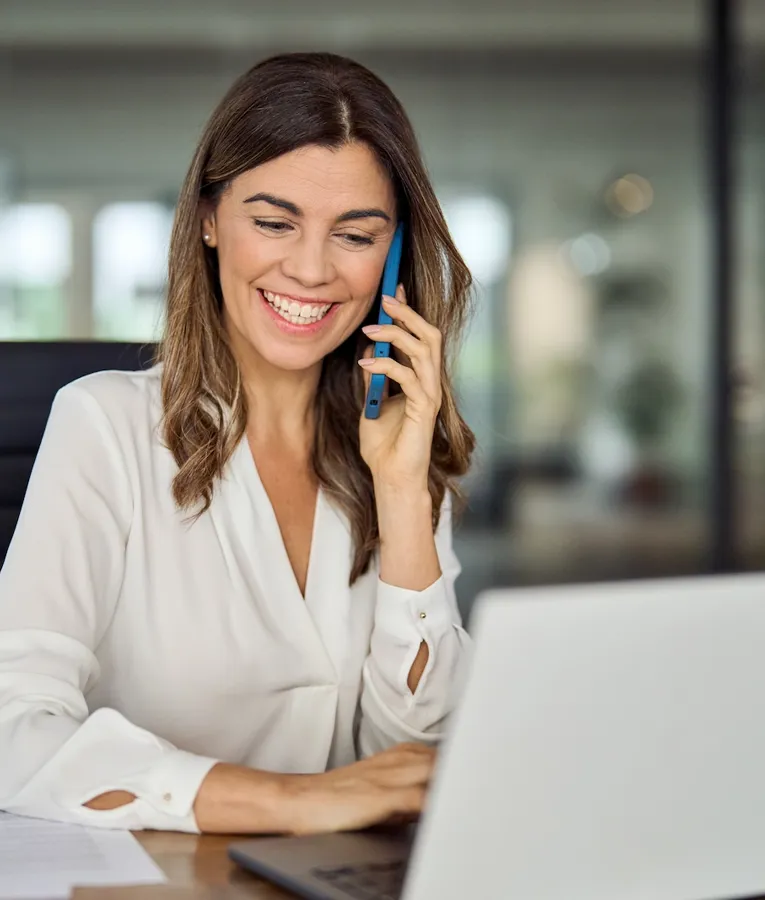Leende affärskvinna pratar i telefon medan hon arbetar på en bärbar dator i ett modernt kontor, vilket representerar effektiv fjärrkommunikation i arbetsstyrkan.